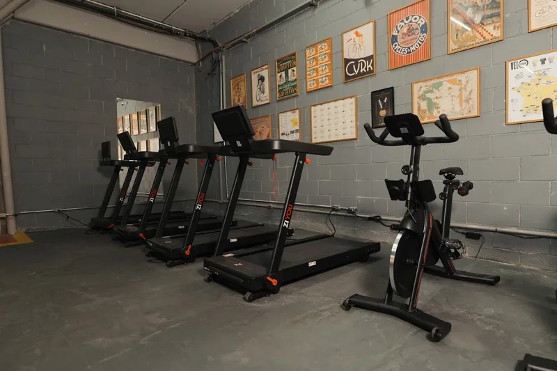 Sala de treino indoor com esteiras e bike, e pôsteres antigos de ciclismo na parede